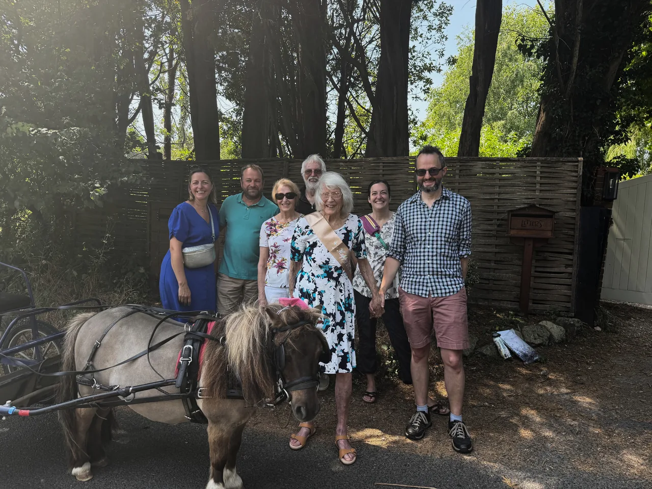 Dot, wearing her birthday sash, stands with her family beside the Shetland pony and chariot on her 100th birthday — her grandchildren travelled from Somerset and the United States to celebrate with her.